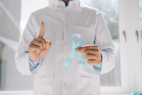 Vista parcial del médico sosteniendo la cinta de conciencia azul y mostrando el gesto de atención - foto de stock