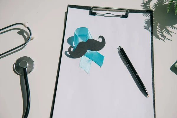 Blue awareness ribbon, paper cut mustache and pen on clipboard with blank paper near stethoscope — Stock Photo