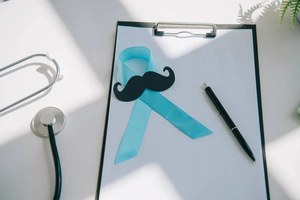 Cinta de conciencia azul, bigote cortado en papel y pluma en el portapapeles con papel en blanco cerca del estetoscopio - foto de stock