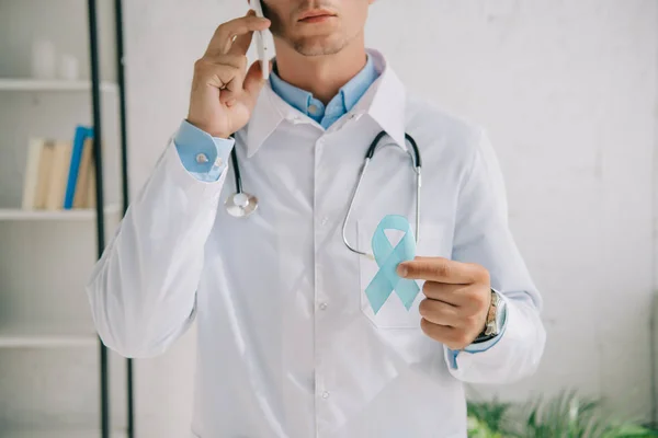 Обрезанный вид доктора, держащего синюю ленту осведомленности во время разговора на смартфоне — стоковое фото