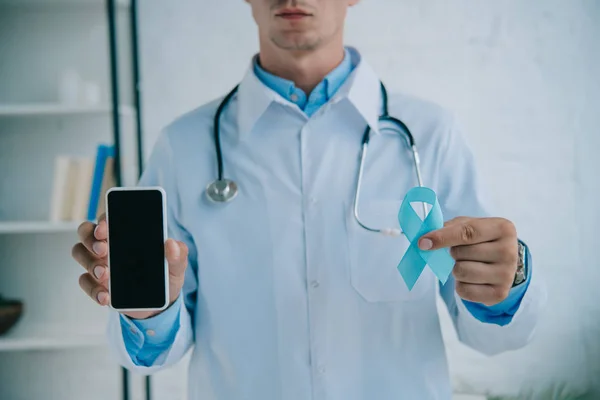 Vue recadrée du médecin tenant un ruban de sensibilisation bleu et un smartphone avec écran vierge — Photo de stock