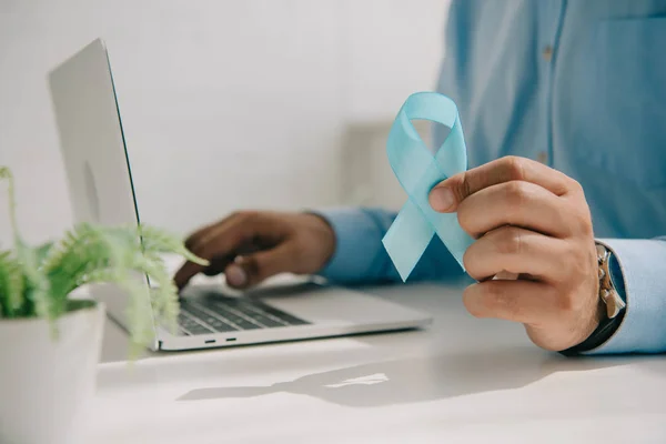 Vista recortada del hombre en camisa azul que sostiene la cinta azul de la conciencia mientras que usa el ordenador portátil - foto de stock