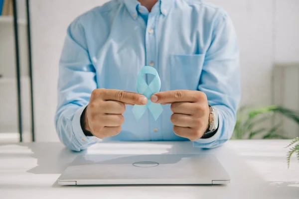 Cropped view of man in blue shirt holding blue awareness ribbon near laptop — Stock Photo