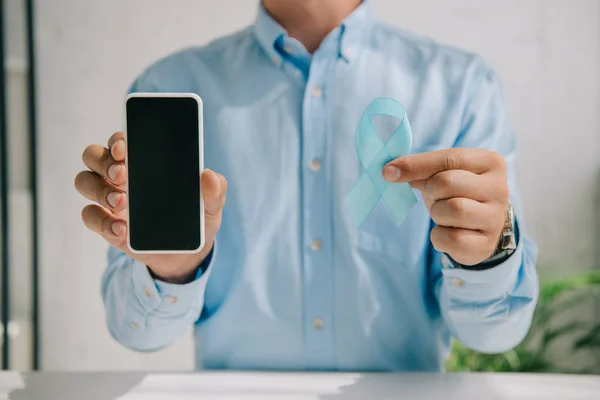 Partial view of man holding blue awareness ribbon and smartphone with blank screen — Stock Photo
