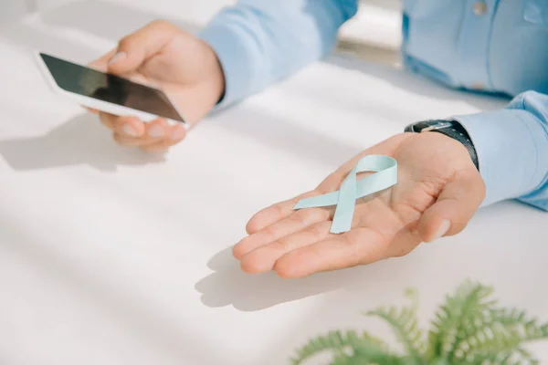 Vista recortada del hombre sosteniendo la cinta de conciencia azul y el teléfono inteligente con pantalla en blanco - foto de stock