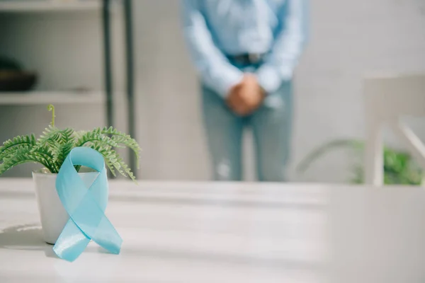 Enfoque selectivo de la cinta de conciencia azul cerca de maceta y el hombre de pie por la pared y sosteniendo las manos apretadas cerca del área de la ingle - foto de stock