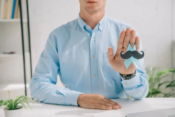 Partial view of man in blue shirt holding blue awareness ribbon with paper cut mustache — Stock Photo