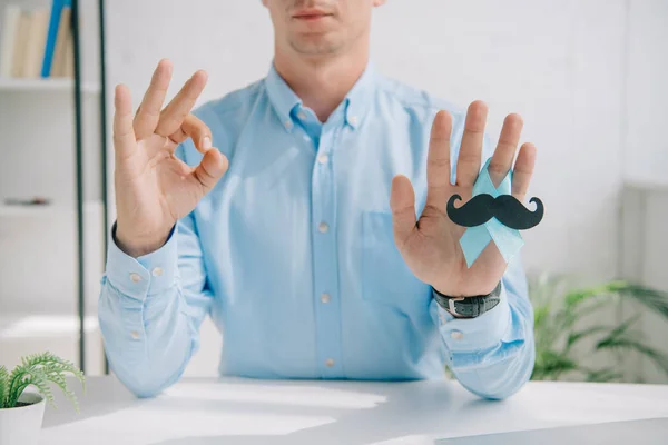 Cropped view of man in blue shirt holding blue awareness ribbon with paper cut mustache and showing ok gesture — Stock Photo