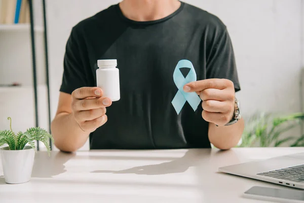 Vue recadrée de l'homme en t-shirt noir tenant ruban de sensibilisation bleu et récipient avec des pilules — Photo de stock