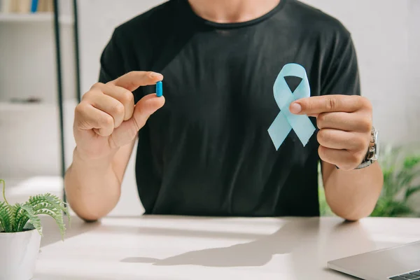 Cropped view of man in black t-shirt holding blue awareness ribbon and pill — Stock Photo