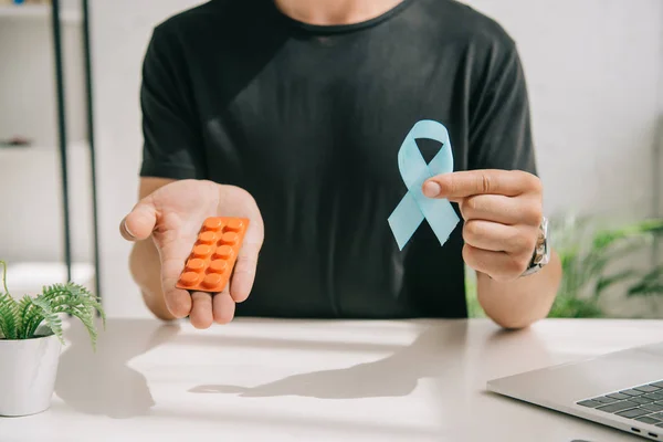 Vue partielle de l'homme en t-shirt noir tenant un ruban de sensibilisation bleu et des pilules — Photo de stock