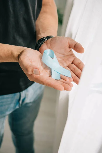 Cropped view of man holding blue awareness ribbon while standing near window — Stock Photo