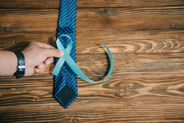 Cropped view of male holding blue awareness ribbon near tie on wooden surface — Stock Photo