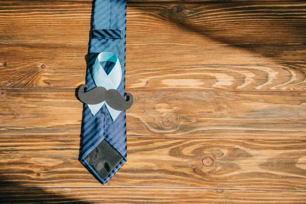 Tie, blue awareness ribbon and paper cut mustache on wooden surface — Stock Photo