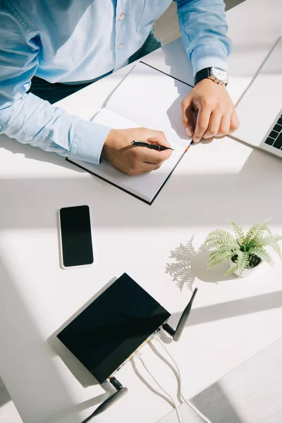 Partial view of businessman writing in notebook near laptop, smartphone and router — Stock Photo