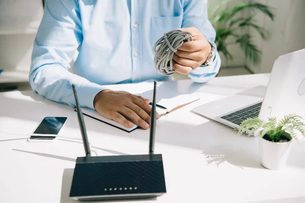 Vista parcial del hombre de negocios sosteniendo el cable cerca de la computadora portátil, teléfono inteligente y router - foto de stock