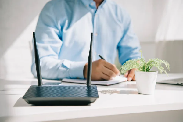 Cropped view of businessman writing in notebook near router and flowerpot — Stock Photo