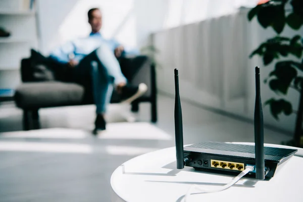 Selective focus of black plugged router on white table and businessman sitting on sofa — Stock Photo