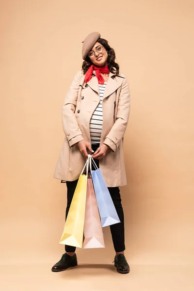 Pregnant french woman in coat holding shopping bags on beige background — Stock Photo