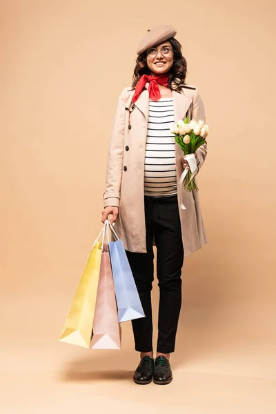 Pregnant french woman in coat holding shopping bags and bouquet on beige background — Stock Photo