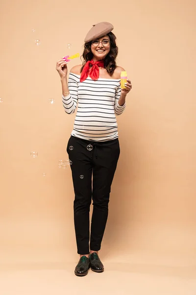 Pregnant french woman in beret holding bottle with soap bubbles on beige background — Stock Photo