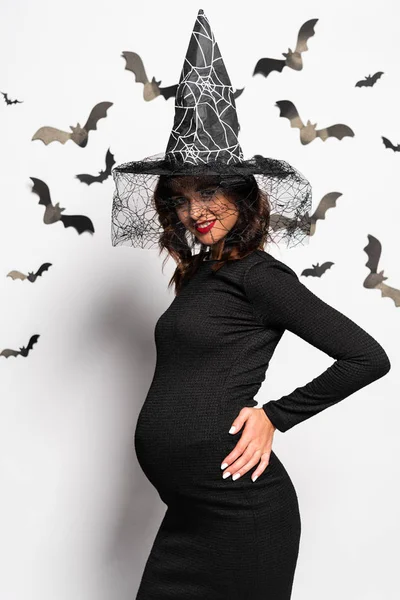 Mujer embarazada en sombrero de bruja sonriendo y mirando a la cámara en Halloween - foto de stock