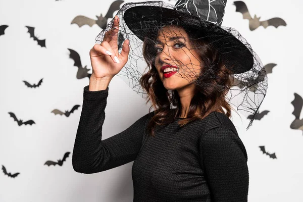 Attrayant femme en chapeau de sorcière souriant et regardant la caméra dans Halloween — Photo de stock