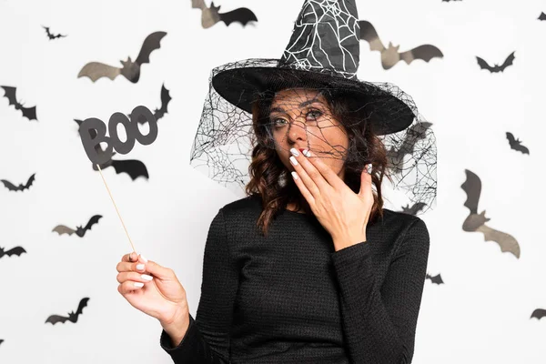 Mujer conmocionada en sombrero de bruja sosteniendo palo con letras boo en Halloween - foto de stock