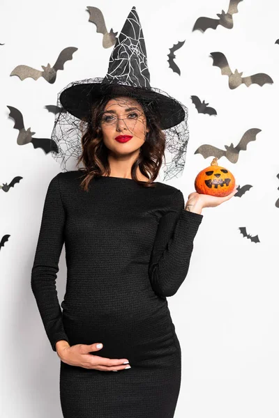 Mujer embarazada con sombrero de bruja sosteniendo calabaza en Halloween - foto de stock