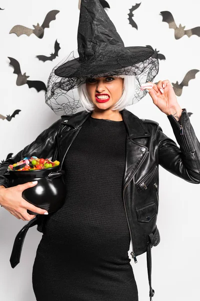 Pregnant and angry woman in witch hat and wig holding pot with candies in Halloween — Stock Photo