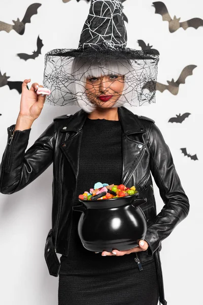 Atractiva mujer en sombrero de bruja y peluca sosteniendo olla con caramelos en Halloween - foto de stock