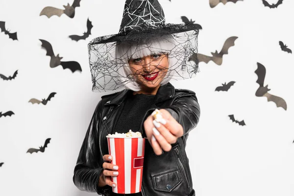 Mujer sonriente con sombrero de bruja y peluca sosteniendo palomitas de maíz en Halloween - foto de stock