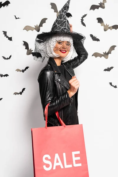 Donna sorridente in cappello da strega e parrucca con shopping bag con lettering in vendita ad Halloween — Foto stock