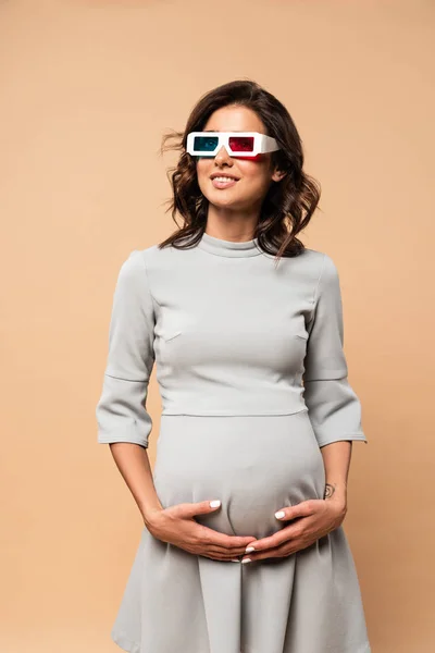 Беременная женщина в сером платье с 3D очки касаясь живота на бежевом фоне — стоковое фото
