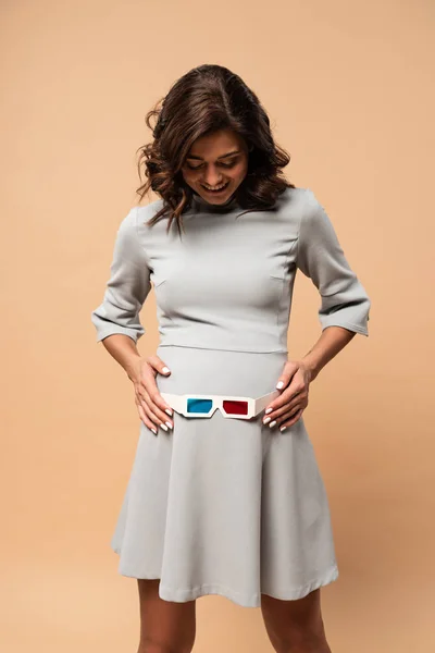 Pregnant woman in grey dress putting on belly 3d glasses on beige background — Stock Photo