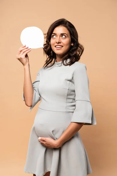 Mulher grávida em vestido cinza tocando barriga e segurando bolha de fala no fundo bege — Fotografia de Stock