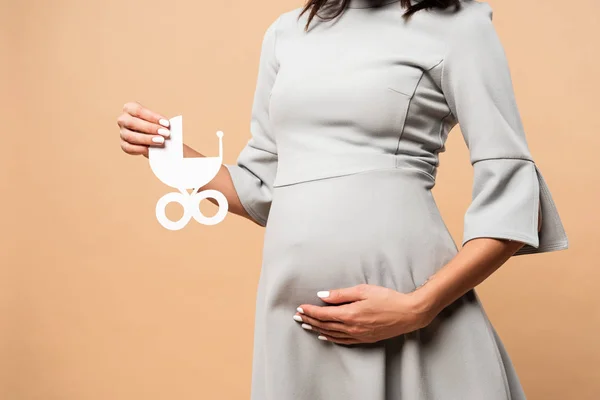 Vista recortada de la mujer embarazada en vestido gris sosteniendo carro de bebé de papel sobre fondo beige - foto de stock
