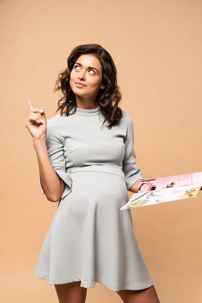 Беременная женщина в сером платье холдинг журнал и показ идеи жест на бежевом фоне — стоковое фото
