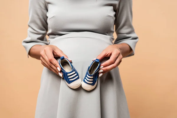 Vista recortada de la mujer embarazada en vestido gris sosteniendo zapatillas de deporte sobre fondo beige - foto de stock