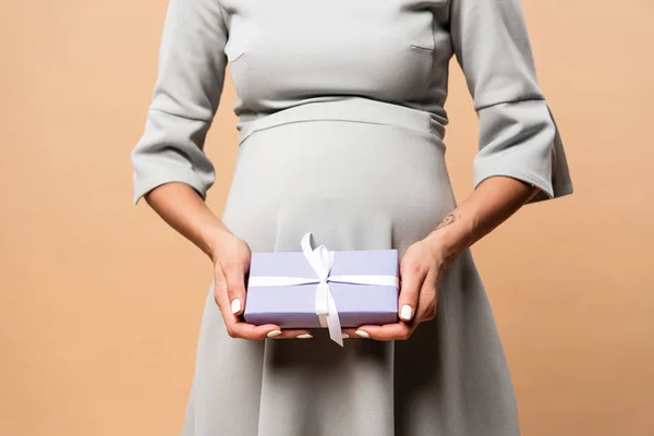Cropped view of pregnant woman in grey dress holding gift on beige background — Stock Photo