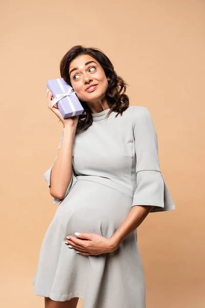 Беременная женщина в сером платье с подарком и трогательным животом на бежевом фоне — стоковое фото