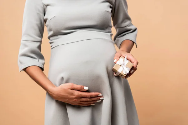 Обрезанный вид беременной женщины в сером платье с подарком на бежевом фоне — стоковое фото