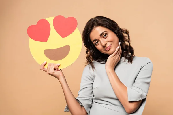 KYIV, UKRAINE - AUGUST 1, 2019: attractive woman in grey dress holding smiley with heart eyes on beige background — Stock Photo