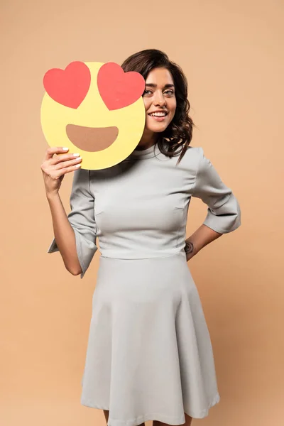 KYIV, UKRAINE - AUGUST 1, 2019: pregnant woman in grey dress holding smiley with heart eyes on beige background — Stock Photo