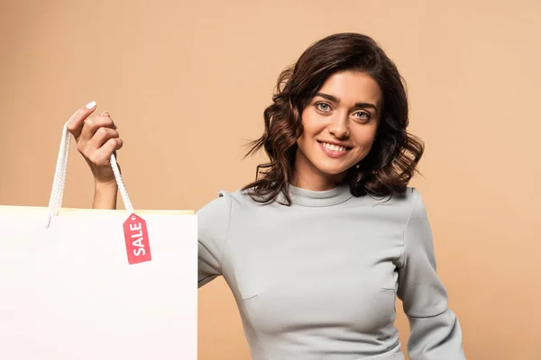 Mujer sonriente en vestido gris sosteniendo bolsa aislada en beige - foto de stock