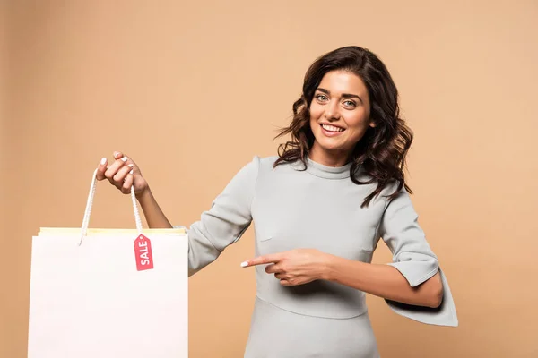 Mujer embarazada en vestido gris apuntando con el dedo a la bolsa de la compra sobre fondo beige - foto de stock