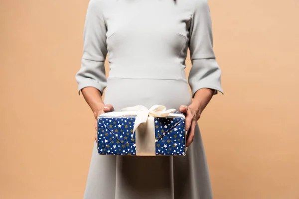 Обрізаний вид вагітної жінки в сірій сукні, що тримає подарунок на бежевому фоні — стокове фото