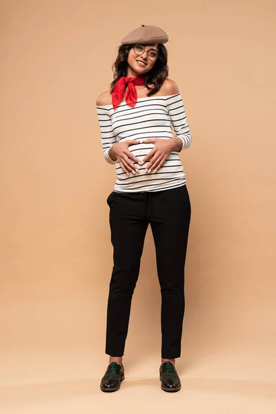 Беременная француженка в берете показывает сердечный жест на бежевом фоне — стоковое фото