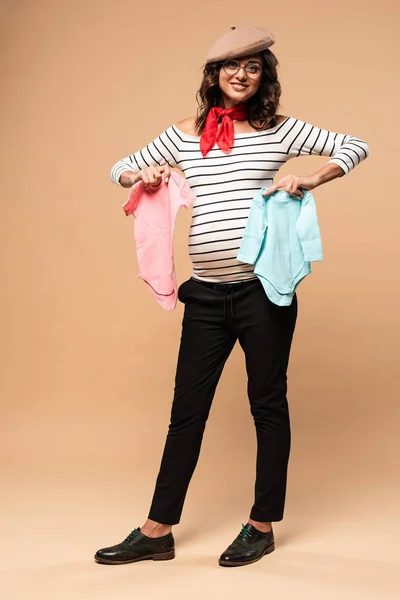 Femme française enceinte en béret tenant des salopettes sur fond beige — Photo de stock