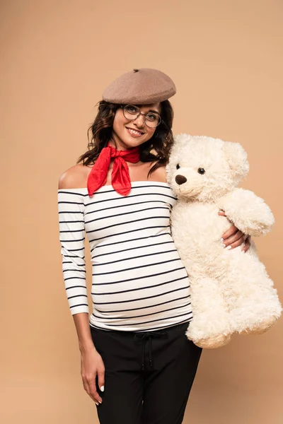 Femme française enceinte en béret souriant et tenant un ours en peluche sur fond beige — Photo de stock